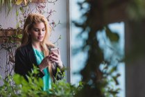 Portrait of blond girl standing at window and browsing phone. — Stock Photo