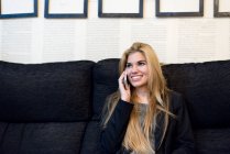 Весела блондинка сидить на дивані і розмовляє по телефону — стокове фото