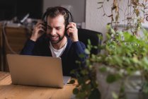 Retrato del hombre sentado en la mesa ajustando los auriculares y mirando hacia abajo portátil - foto de stock