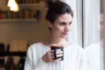 Portrait of brunette girl posing with cup near window and looking aside — Stock Photo