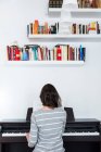 Visão traseira da menina tocando piano perto da parede com prateleiras de livros — Fotografia de Stock