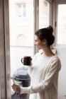 Вид сбоку девушки-брюнетки с чашкой в руках, открывающей окно дома — стоковое фото