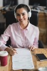 Portrait of smiling worker in headphones looking at camera and signing papers at workplace — Stock Photo