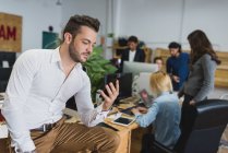 Portrait of man sitting on table and using phone over office workers on backdrop — Stock Photo