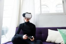 Жінка в окулярах VR тримає геймпад і сидить на дивані. Горизонтальний постріл у приміщенні — стокове фото