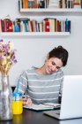 Lächelnde Frau sitzt am Laptop und blickt auf Notizbuch herab — Stockfoto