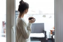 Вид збоку дівчини в білому светрі, що позує біля вікна з чашкою в руках — стокове фото
