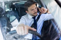 Молодой человек в костюме разговаривает по телефону во время вождения автомобиля — стоковое фото