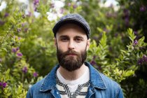 Portrait of bearded man wearing cap and denim jacket looking unemotionally at camera in park. — Stock Photo