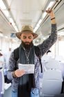 Portrait of bearded man in hat standing in train and looking down at map in hands — Stock Photo