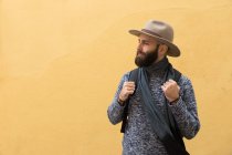 Bearded man with backpack posing over yellow wall and looking away. — Stock Photo