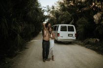 Retrato de pareja con rastas abrazándose en camino de bosque tropical con furgoneta estacionada - foto de stock