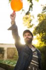 Cheerful bearded man in cap holding orange balloon above head and smiling in sunlight. — Stock Photo