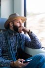 Задумчивый бородатый мужчина сидит в поезде и слушает музыку с наушниками — стоковое фото