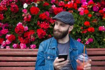 Sonriente hombre barbudo sentado en el banco junto a las flores con batido y teléfono inteligente en las manos y mirando hacia otro lado - foto de stock