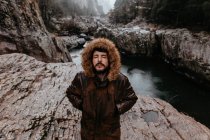 Бородатий чоловік стоїть з закритими очима і руками в кишенях на тлі гірської річки — стокове фото