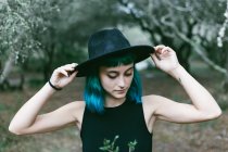 Close up portrait of stylish girl with short blue hair toughing her hat while standing in park. — Stock Photo