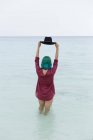 Неузнаваемая сексуальная девушка в красной рубашке, стоящая в море и держащая черную шляпу над своей голубой головой. — стоковое фото