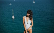 Portrait of brunette woman wearing white swimsuit looking to camera and posing against ocean with floating yachts — Stock Photo