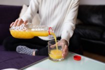 Mid section of female sitting on coach and pouring orange juice in glass on table — Stock Photo