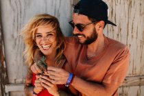 Portrait of cheerful blonde girlfriend laughing at camera while hugging with her bearded boyfriend in cap and sunglasses — Stock Photo