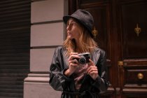Молодая женщина в кожаной куртке и шляпе держит камеру и смотрит в сторону — стоковое фото