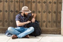Bearded man in earphones sitting on ground and searching in backpack. — Stock Photo