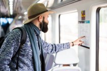 Homem barbudo de chapéu apontando com o dedo no mapa da estação em vagão de trem — Fotografia de Stock