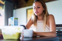 Jovem mulher sentada e navegando smartphone enquanto toma café da manhã na cozinha . — Fotografia de Stock