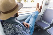 Side view of bearded man in hat putting legs on window and relaxing in train. — Stock Photo