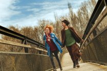 Dos mujeres corriendo por el puente tomadas de la mano. Horizontal al aire libre tiro. - foto de stock