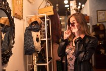 Junge stylische Frau probiert Sonnenbrille vor Spiegel in Accessoires-Geschäft an. — Stockfoto