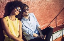 Interracial couple in sunglasses sitting by wall and looking at camera — Stock Photo