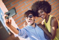 Cheerful interracial couple taking selfie against brickwall — Stock Photo