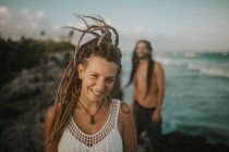 Портрет улыбающейся девушки с дредами, смотрящей в камеру на мужчину, позирующего на галечном тропическом пляже — стоковое фото