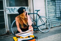 Mujer con gorra sentada en la calle con papeles hablando por teléfono cerca de la bicicleta. - foto de stock