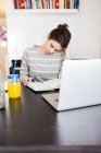 Brunette fille assise à table avec ordinateur portable et pot de jus d'orange et regardant vers le bas au carnet — Photo de stock
