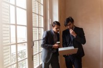 Двоє молодих бізнесменів стоять біля вікна і дивляться на ноутбук . — стокове фото