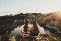 Rückansicht von zwei Mädchen, die in amerikanische Flagge gehüllt an einem abgelegenen Bergsee sitzen — Stockfoto