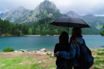 Girls under umbrella standing at mountain lake shore — Stock Photo