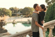 Portrait of boyfriend embracing girlfriend and kissing in cheek on stairs in park over lake on backdrop — Stock Photo