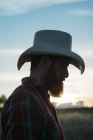 Vue latérale de l'homme barbu en chapeau de cow-boy posant à la campagne au crépuscule — Photo de stock
