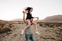 Glückliches junges Paar lacht und lächelt in der Landschaft vor Bergen und blauem Himmel. Mann hält Frau auf Schultern. — Stockfoto