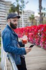 Вид сбоку бородатый мужчина опирается на перила с смузи в руке и просматривает смартфон — стоковое фото