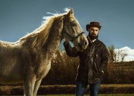 Бородатый мужчина держит лошадь и смотрит в камеру на сельскую местность — стоковое фото