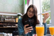 Portrait of brunette woman in eyeglasses holding toast and browsing smartphone at cafe table — Stock Photo