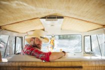 Man in cowboy hat sitting at drivers seat of retro van and looking over shoulder at camera — Stock Photo
