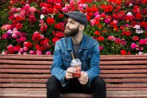 Porträt eines bärtigen Mannes auf einer Bank sitzend mit Smoothie auf dem Hintergrund von Rosensträuchern — Stockfoto