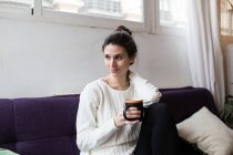Portrait of brunette girl posing on coach with cup of coffee and looking aside — Stock Photo