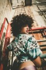 Expressive jeune fille avec afro regardant par-dessus l'épaule à la caméra sur la scène industrielle — Photo de stock